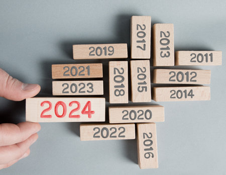 Photo of wooden blocks with gray numbers and one of them standing out and hold by a human hand with red number 2024 imprinted on wooden surface. Gray Background. New Year, Future, Vision and Standing Out concept.の写真素材