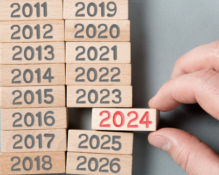 Photo of wooden block rows with gray numbers and one of them held by a human hand with red number 2024 imprinted on wooden surface. Gray background. New Year concept.の写真素材