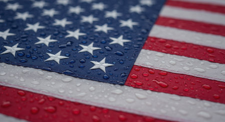 A poignant close-up photograph of an American flag covered in shimmering raindrops. The water droplets highlight the vibrant colors and fabric texture, creating powerful metaphors for both sorrow and remembrance (like tears) as well as resilience and endurance through storms. This meaningful image is suitable for Memorial Day, themes of national mourning, or symbolizing the nation's strength and perseverance through difficult times.の素材