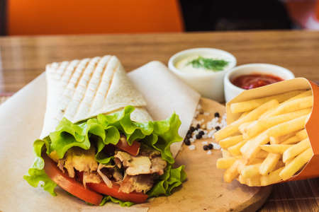 Shawarma with chicken meat and french fries in a box on a table in a cafe. Burrito on a wooden board...