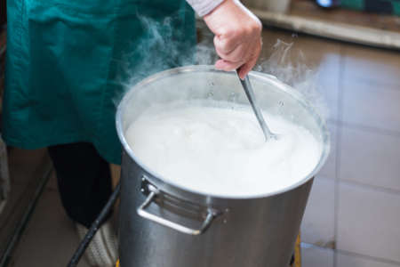 A woman cooks milk in a vat and makes cottage cheese and cheese at home. Production of cheese on the farm. Aluminum container with boiling milk and steam over it.の写真素材