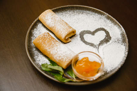 pancakes rolls on a dark plate with powdered sugar, jam and mint. Delicious dessert and a heart of...
