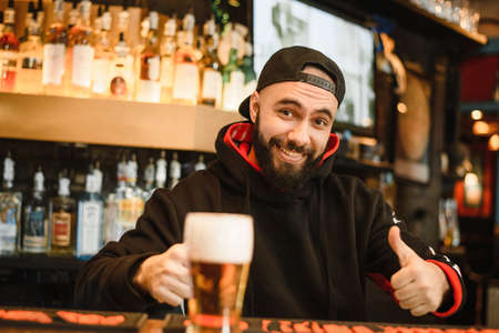 Smiling bearded bartender gives beer in a glass. Friendly man working at the bar.