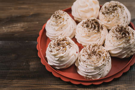 Cream-colored Pavlova meringue lies on a red tray on a wooden old table. Chocolate coated meringue sweets recipe on a brown background.の写真素材