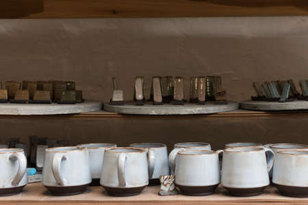 glazed ceramic dishes stand on a shelf in the workshop shelving with handmade ceramic and clay mugs.