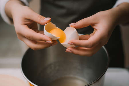 Female cook in a gray apron breaks an egg in a metal bowl. Separate the protein from the yolk.の写真素材