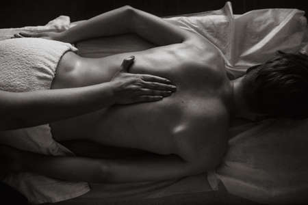 Woman massages under the shoulder blade on the back. Masseur does massage to a man lying on a couch.