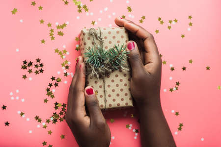 Black girl is holding a gift of Kraft paper on a pink background with confetti. Womans hands holding...