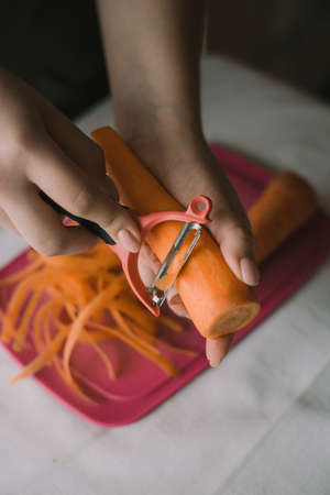 Cook peels carrots in the kitchen with a special knife. The girl peels orange carrots with a peeler.