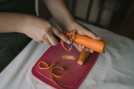 Cook peels carrots in the kitchen with a special knife. The girl peels orange carrots with a peeler.の写真素材