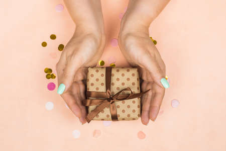 Hands of girl with blue pastel manicure holding craft gift with bow on pink background with ....
