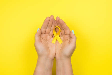 Yellow gold ribbon in the hands of a girl on a yellow background. Children cancer concept. Awareness month. Childhood Cancer Day February, 15.の写真素材
