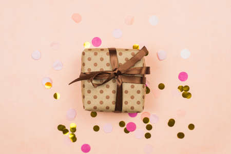 Gift in craft packaging with brown satin ribbon on pastel pink background with gold and pink confetti. Gift giving concept, a place for your text and advertising. Concept of Christmas, happy New year, Birthday and different holidays.の写真素材