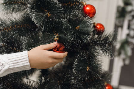 A hand in a white knitted sweater hangs a red Christmas ball on the branch of an artificial Christmas tree. A close shot of decorating the Christmas tree with toysの写真素材