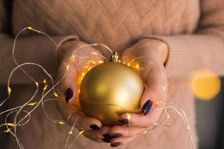 A girl in a pink sweater holds and gives a golden Christmas ball in her hands with a yellow garland and lights. Beautiful tinted. The concept of Christmas, New Year, warmth, celebration.の写真素材