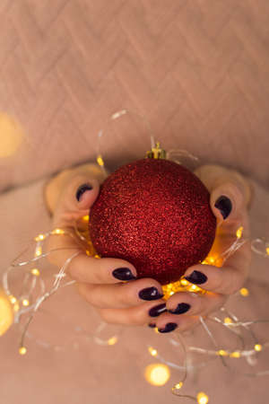A girl in a pink sweater holds and gives a red shiny Christmas ball in her hands with a yellow garland and lights. Beautiful tinted. The concept of Christmas, New Year, warmth, celebration.の写真素材