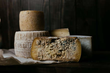 Different homemade cheeses on a dark wooden background. Pieces and cheese heads of Gorgonzola, Asiago, Maasdam, Gauda, Paramezan and Edam. Goat and cow cheese on baking paper. Handwork. Close up.の写真素材