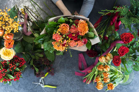 The concept of gifts and bouquets for March 8 and Mother's Day. Florist creates a bouquet in a flower shop. Top view of a beautiful bouquet of red, orange, burgundy, yellow roses, tulips. View from above. Flat lay composition.の写真素材