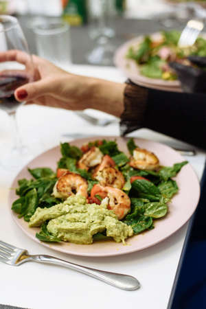 Fried prawns on a dish with spinach and guacamole pasta salad. The girl drinks red wine from a glass and eats an expensive dish in a restaurant.の写真素材