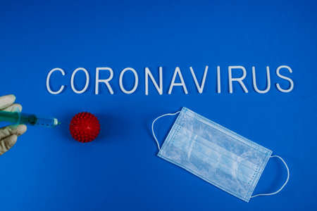A doctor in a plastic white glove injects a syringe into a red ball of coronavirus. Treating infection. The word coronavirus and mask for the respiratory tract on a blue background. Flat lay.の写真素材