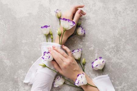 Tender hands of a girl with nude manicure on a gray grunge background with eustoma flowers. Creative illustration of hand skin care, natural cosmetics components. Hands of a cute girl with light white sleeves with flowering plants.の写真素材