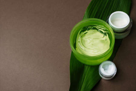 Jars of moisturizing white cream and green aloe gel on a palm leaf. Different moisturizers for different areas of the face on a brown background with copyspace.の写真素材