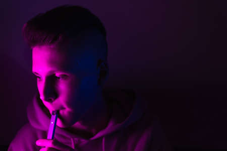 A young guy smokes an electronic cigarette in neon light. Teenage boy soars vape in the studio.の写真素材