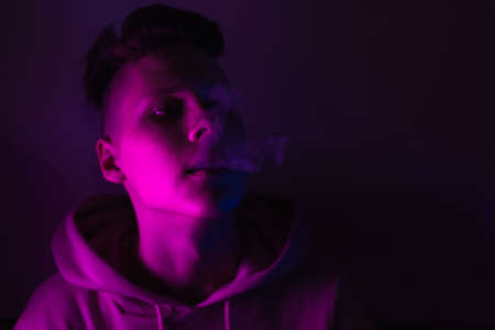 A young guy smokes cigarettes in a neon light. The problem of drug trafficking among adolescents....