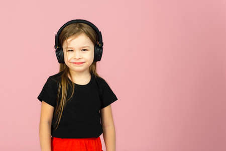 A little girl in wireless big black headphones on a pink pastel background looks at the camera. The...