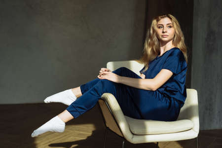 A beautiful girl sits in a comfortable chair in the middle of the room in the rays of the rising sun. The concept of quarantine, self isolation and stay home.の写真素材