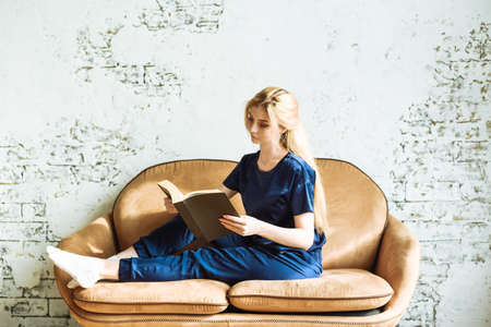 A young girl sits at home on a beige sofa and reads a book. Concept stay home.の写真素材