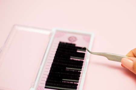 Girl holds tweezers over a pink box with black eyelashes on a pastel background. Eyelash Extension tools.の写真素材