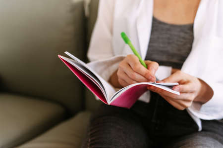 A young girl writes thoughts in her diary. A pretty woman sits on a sofa at home and writes in a notebook on her lap.の写真素材