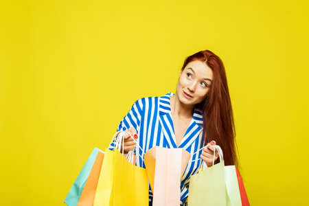 Beautiful woman on a yellow background looks in bags with clothes and different purchases after shopping and is surprised.の写真素材
