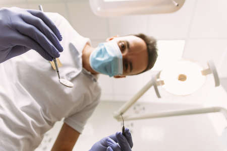 Professional dental doctor is working in office. Young man dentist in protection gloves and a mask, keeps dental instrument looking at the camera.の写真素材