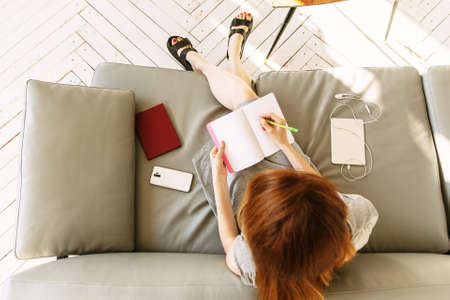 Overhead Shot Looking Down On Woman Working At Notebook At Home Lying On Sofa. A woman writes in a...