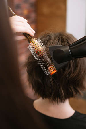 The hairdresser does styling for short hair with a hairdryer and round bristles. Woman doing...