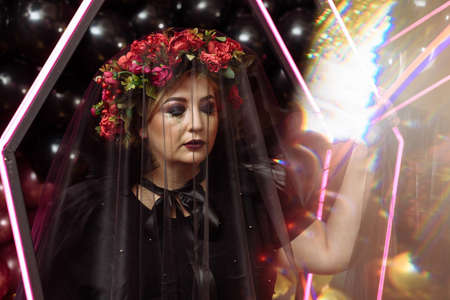 A scary adult witch in a neon coffin covered with a dark veil. Halloween party.