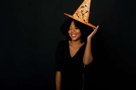 Horizontal shot of a joyful young black woman with afro haircut wearing a Halloween orange hat and black dress. Studio portrait on black background.の写真素材