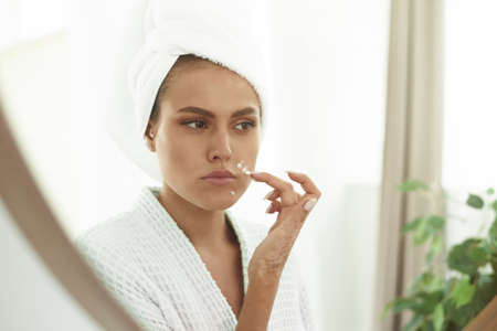 Portrait of a young smiling woman applying a cream for problem skin to her face in the bathroom. A...