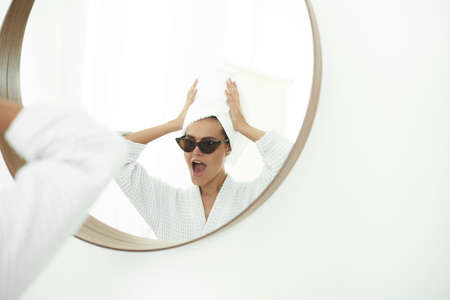 Young woman in bathrobe, with a white towel on her head and black sunglasses looking in bathroom mirror.の写真素材