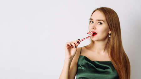 Cute young woman in green silk Christmas dress is biting a candy cane on a white background.