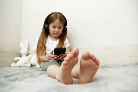 The girl sits in big headphones and looks at the smartphone screen, sitting on her bed.の写真素材
