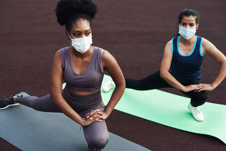 Two different ethnic girls stretch, stretch their muscles before training outdoors in a protective mask. Prevention of coronavirus.の写真素材