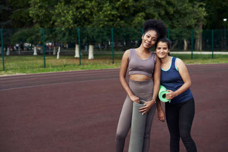 Two sports women hold a yoga mat in their hands and go to train at the stadium on the street. Beautiful models during outdoor workout.の写真素材