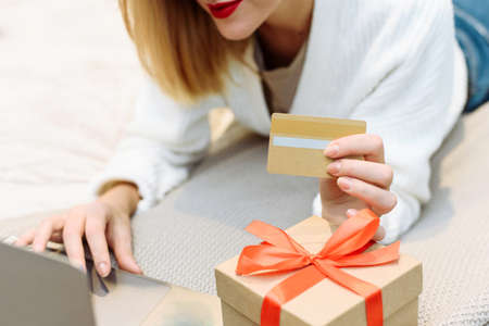 Woman buy presents, prepare to xmas, nearby gift box. Women wearing white sweater lying on the sofa and using laptop and credit card to shopping online at home. Winter holidays sales.の写真素材