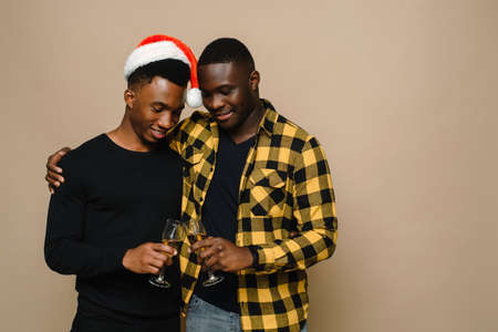 Happy family portrait on Christmas, gay male couple on beige background. A pair of black men in love clink glasses of champagne. Cheers. Celebrate Christmas and New Year.の写真素材