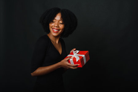 A black woman in a festive black dress holds a Christmas red present with a white ribbon in her hands. The concept of giving or receiving gifts for the new year.の写真素材