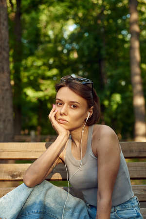 Fashionable stylish young woman listening to music on wired headphones while sitting on a park...