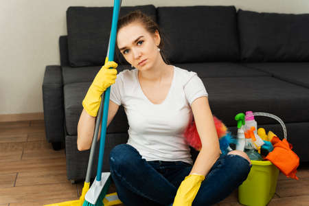A tired housewife sits on the floor and sighs and gets angry. Fatigue from home routine. A woman in rubber gloves next to a coat and cleaning products.の写真素材